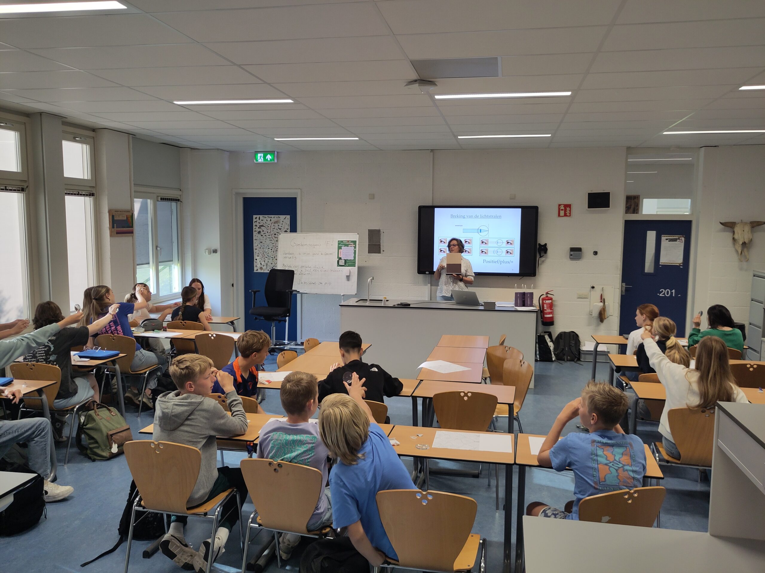 Een leerkracht staat vooraan bij een kleinschalig onderwijs in Middelbare school Westland en spreekt leerlingen toe die met opgestoken handen aan een tafel zitten. Achter de leerkracht wordt een presentatie getoond op een scherm. | ISW Sweelincklaan