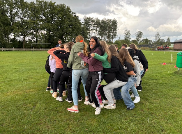 Een groep tieners staat in een krappe cirkel op een grasveld, leunt op elkaar en lacht tijdens een teambuildingactiviteit in de buitenlucht; bomen en een bewolkte lucht op de achtergrond. | ISW Sweelincklaan
